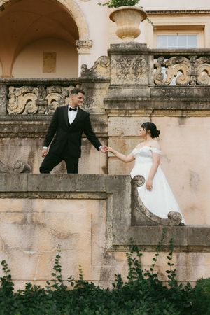 A couple in wedding attire playfully runs along a historic stone wall with greenery at its base.