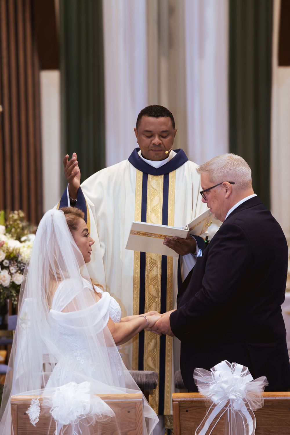 Bride and groom exchange vows at church altar as priest performs Catholic wedding ceremony blessing.