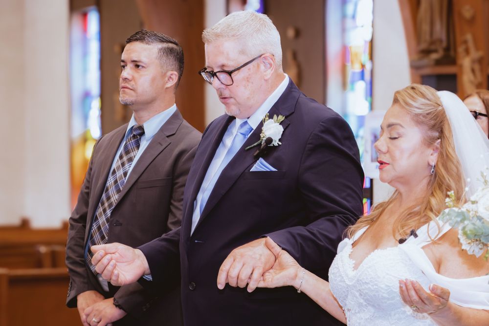 Wedding party stands together in church aisle holding hands during Catholic marriage ceremony prayer.