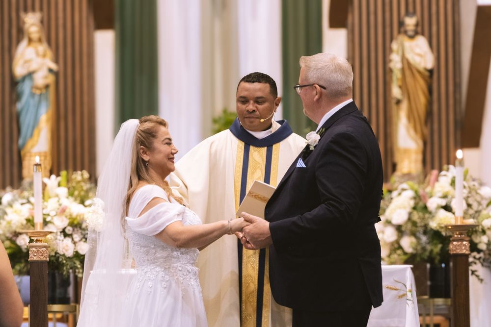Smiling bride and groom hold hands at church altar during Catholic wedding ceremony with priest.