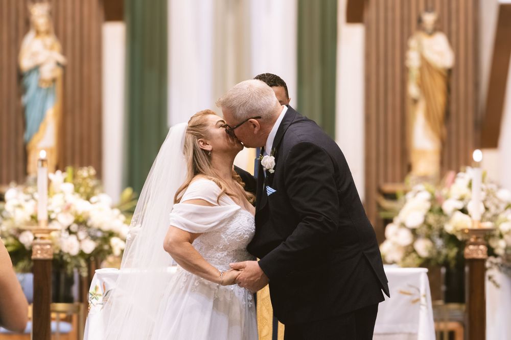 Bride and groom share first kiss at decorated church altar during traditional Catholic wedding ceremony.