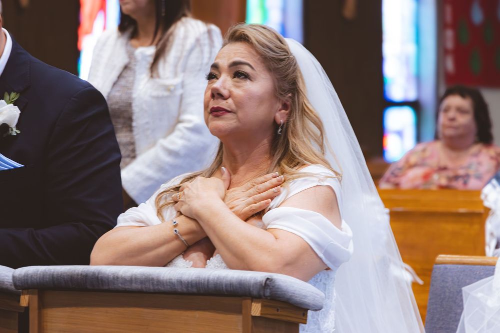 Emotional bride in white wedding dress and veil kneels in prayer during church wedding ceremony.