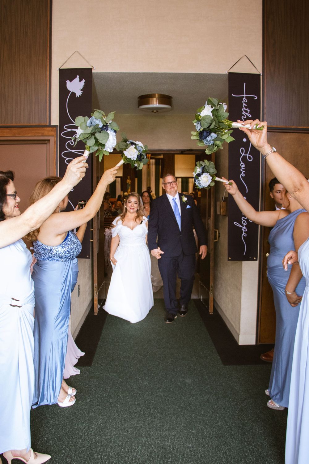 Newlyweds walk through arch of raised bouquets held by bridesmaids in blue dresses after ceremony.