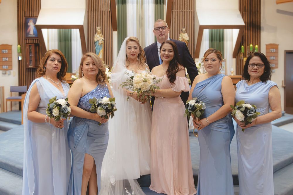 Bride poses with five bridesmaids in blue dresses holding white and blue bouquets inside church.