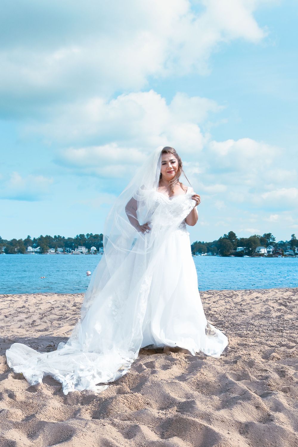 Bride in white wedding gown and long veil poses gracefully on sandy beach by waterfront.
