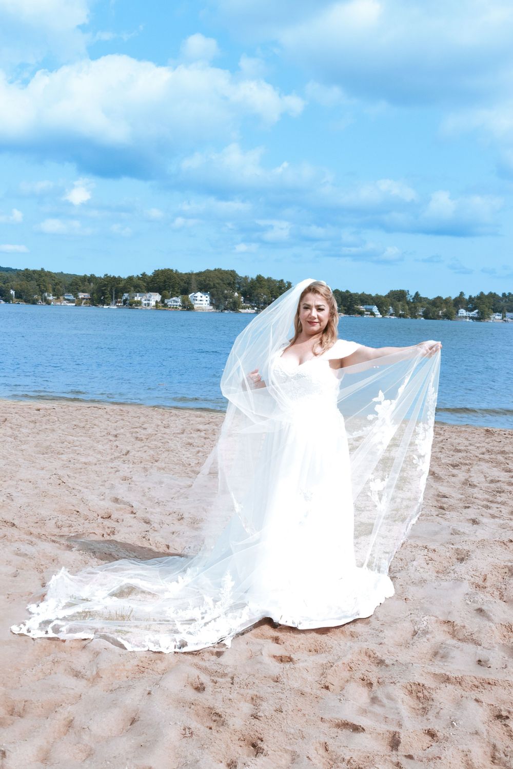Bride in white gown and veil posing on sandy beach with blue sky and lake in background.