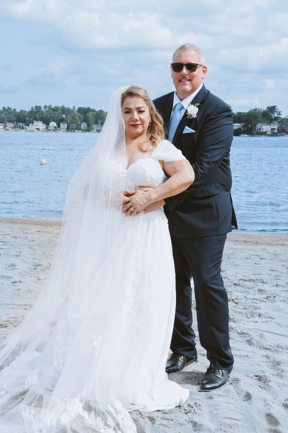 Bride and groom posing together on sandy beach with lake and trees in background on wedding day.