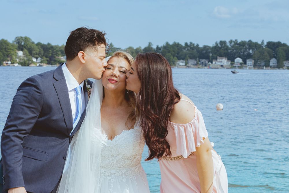 Bride receiving kisses on cheeks from two guests near waterfront on her wedding day.