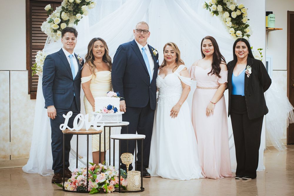 Wedding party group photo with bride, groom, and family members near cake table.
