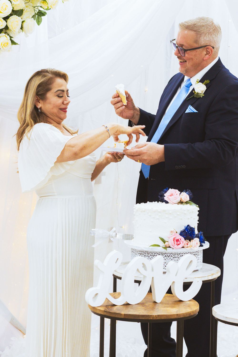 Bride and groom cutting white wedding cake decorated with flowers and Love sign.