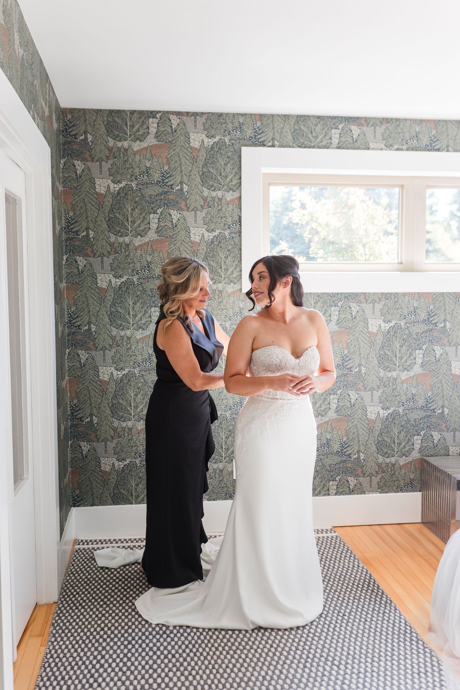 A bride in a white wedding dress gets help with final adjustments in a room with geometric wallpaper.
