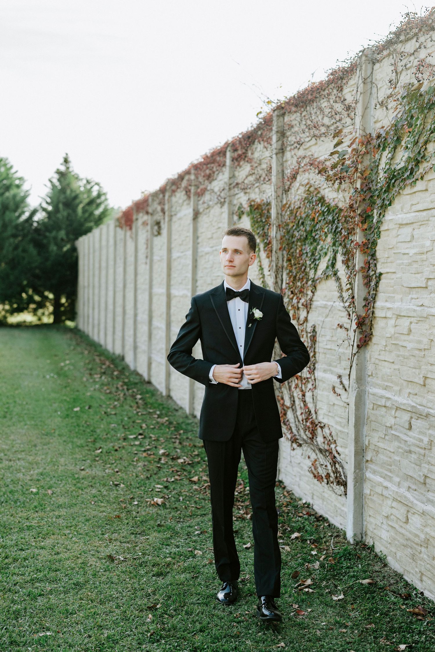 A person in a classic black tuxedo and bowtie stands elegantly against a vine-covered stone wall on a grassy lawn.