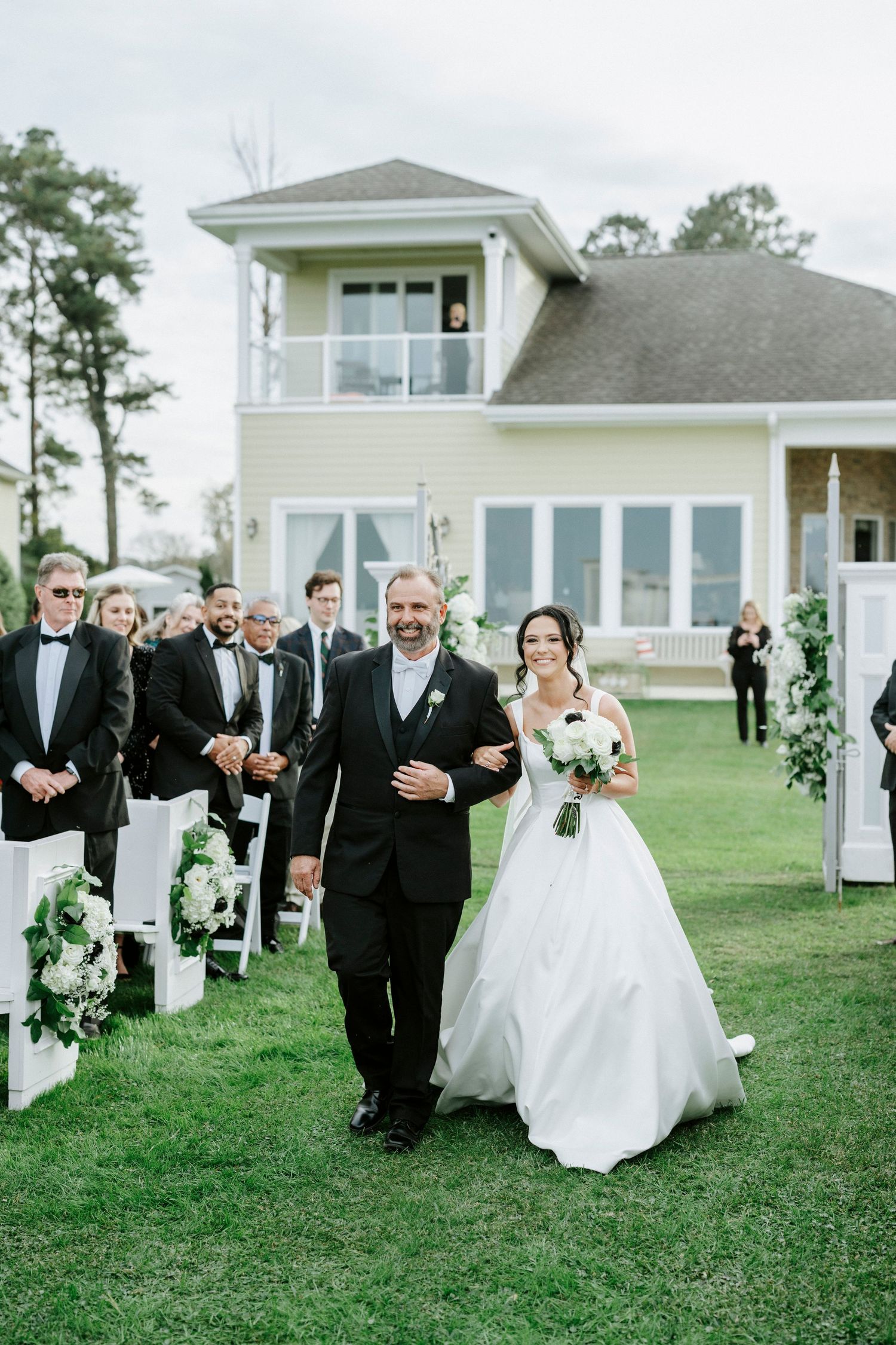 Wedding party walks across a green lawn outside a white coastal home during an elegant outdoor ceremony.