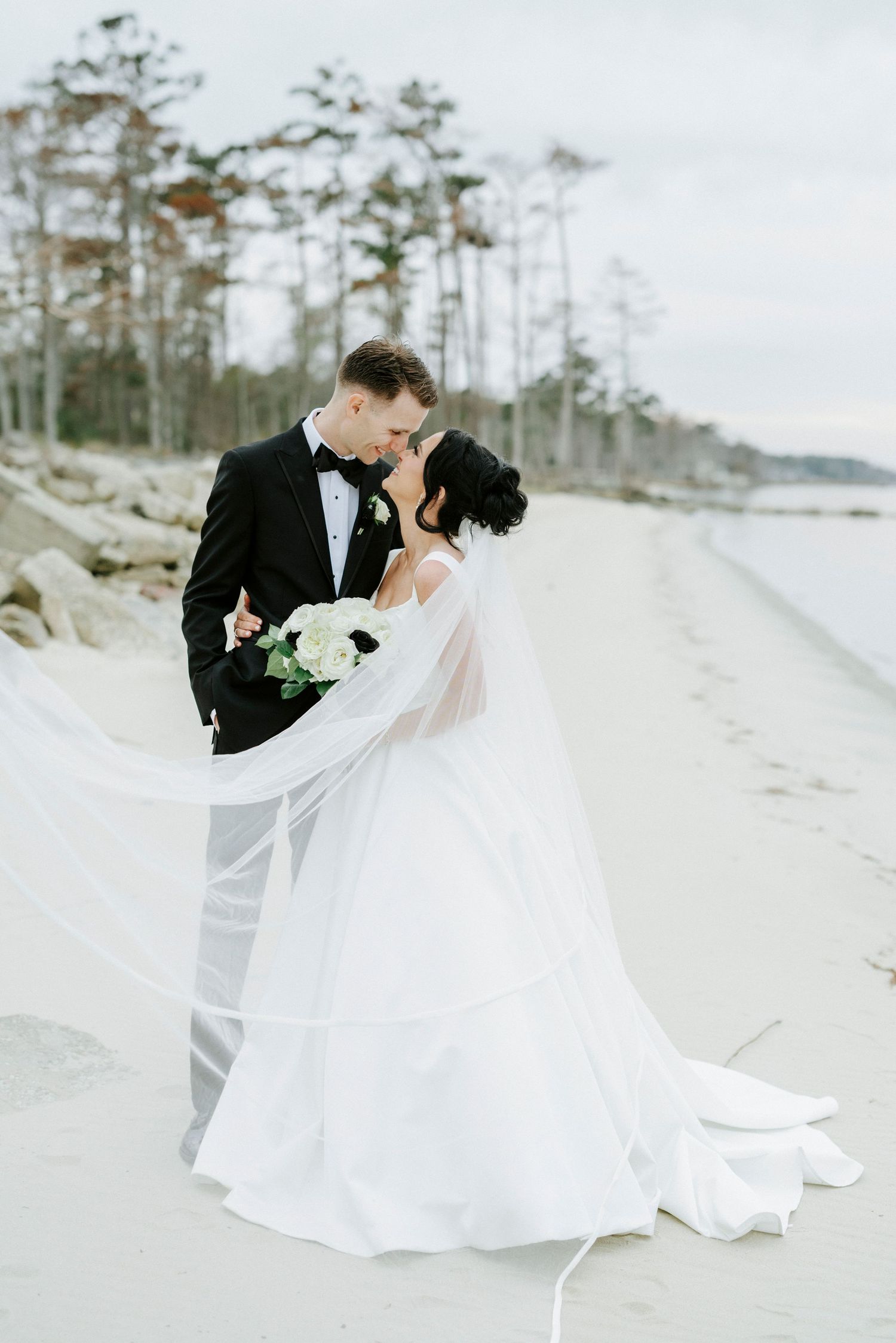 A couple in wedding attire shares a romantic moment on a scenic beach with pine trees in the background.