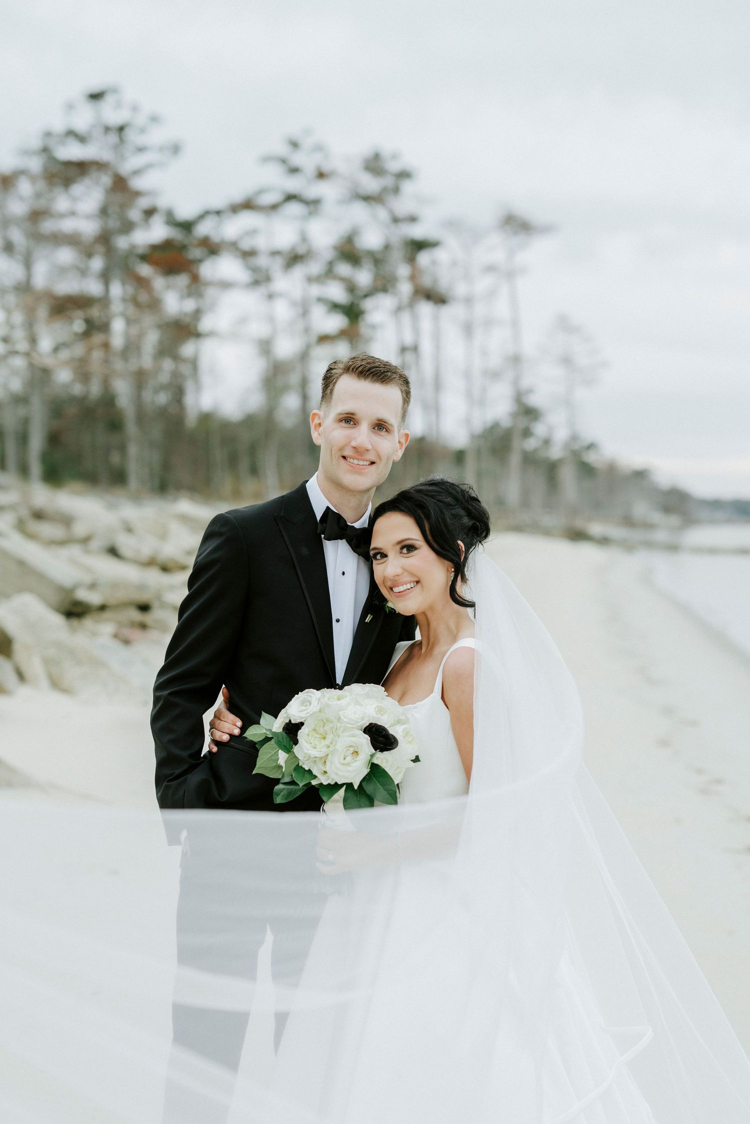 A couple in wedding attire poses together on a serene beach with pine trees in the background.