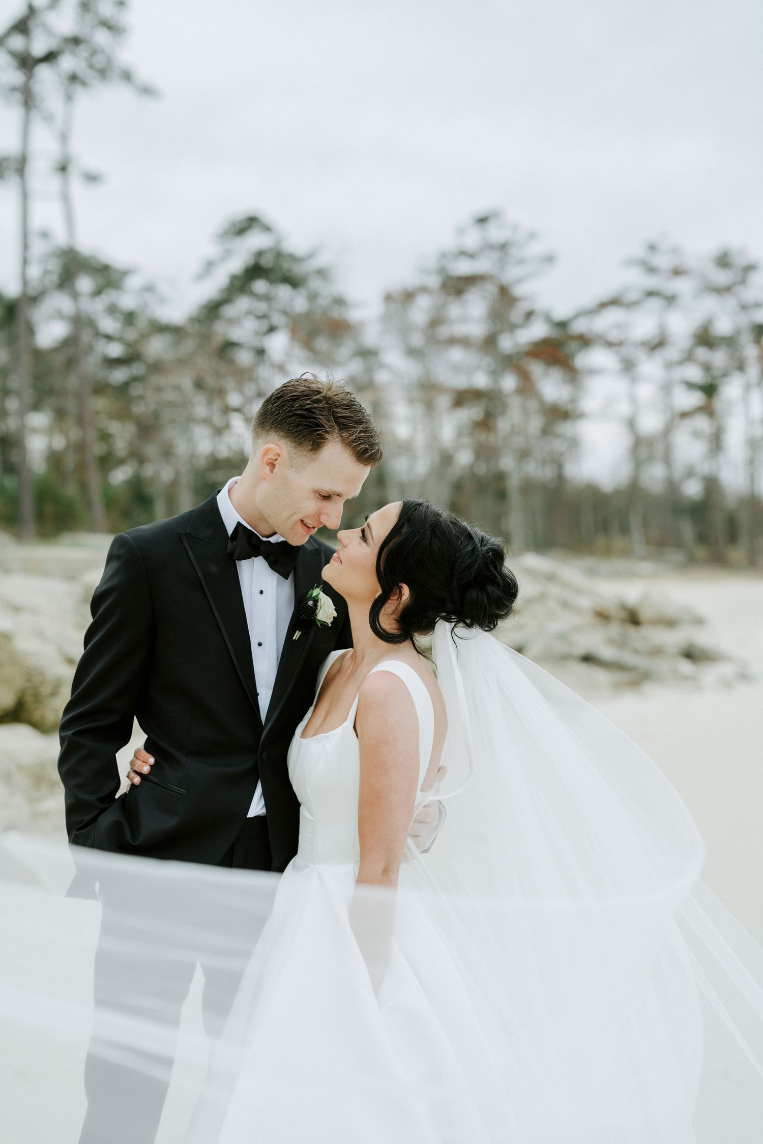 Romantic wedding couple sharing an intimate moment on a beach with pine trees in the background.