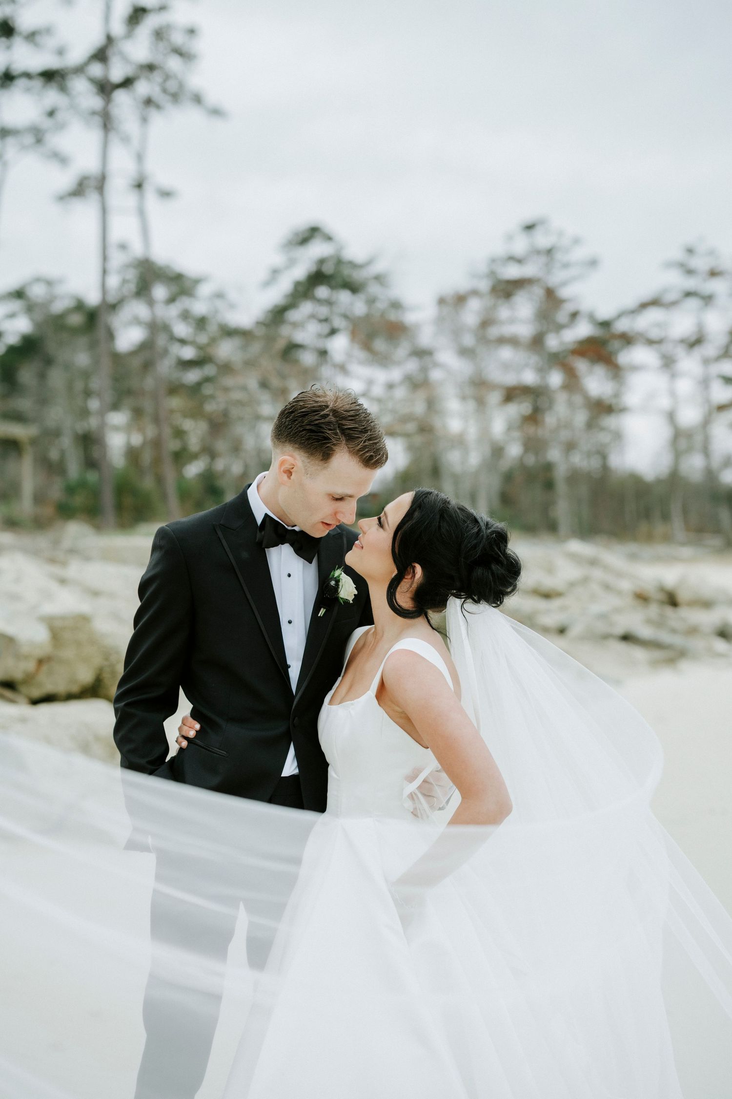 Newlyweds in black and white attire embrace on a cloudy beach with flowing veil.