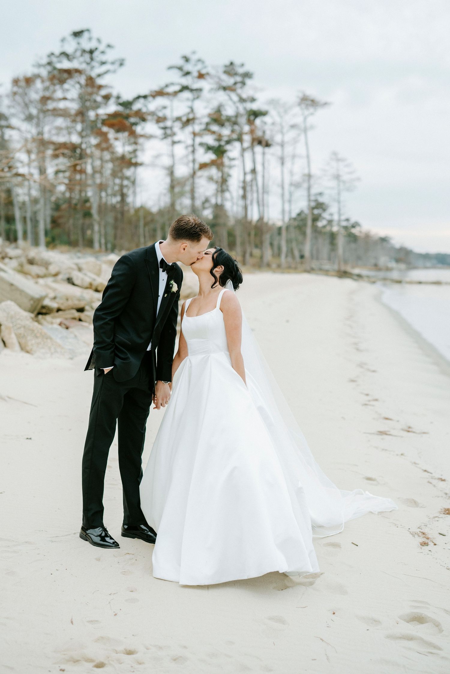 Wedding couple shares tender moments on sandy beach with pine forest backdrop.