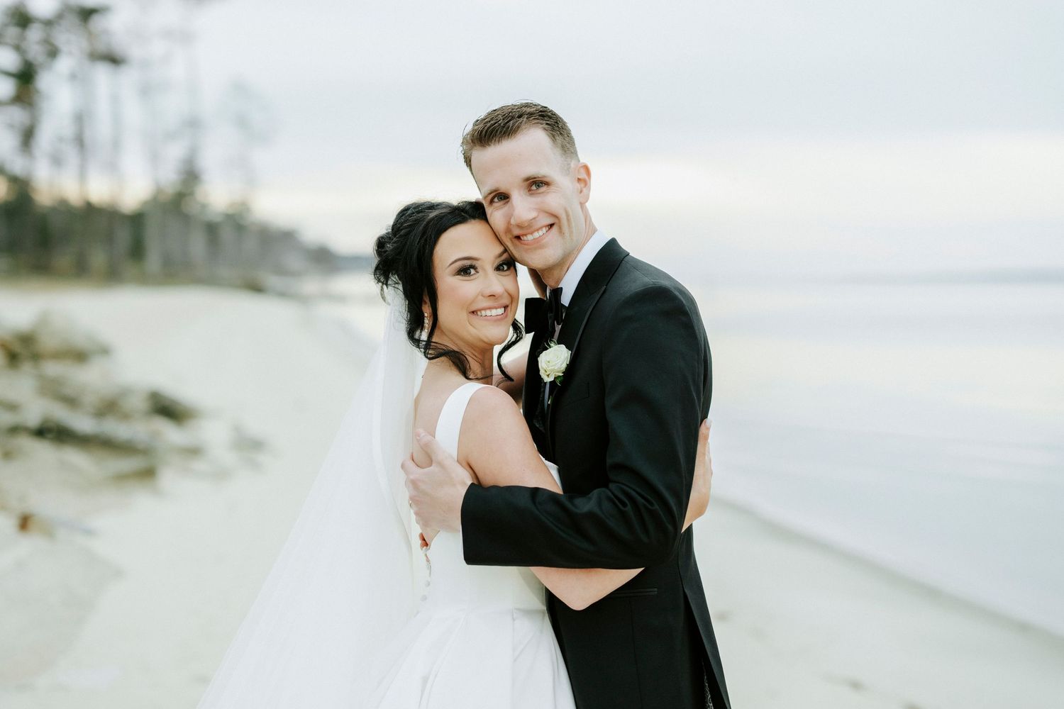 A couple in wedding attire embrace on a serene beach at dusk with the ocean and coastline in the background.