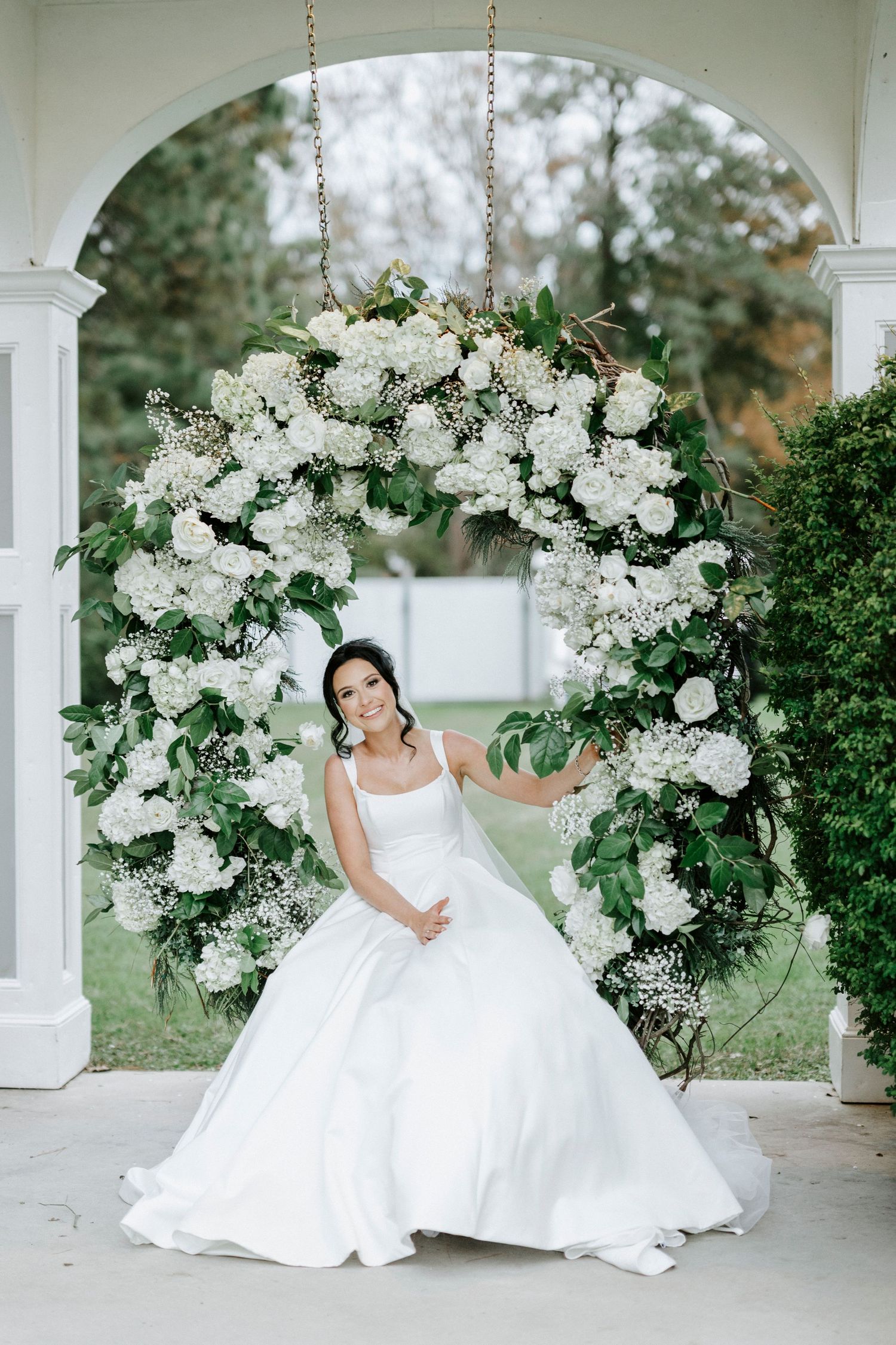 A white floral arch adorns an elegant porch setting with a bride in a flowing white ballgown.