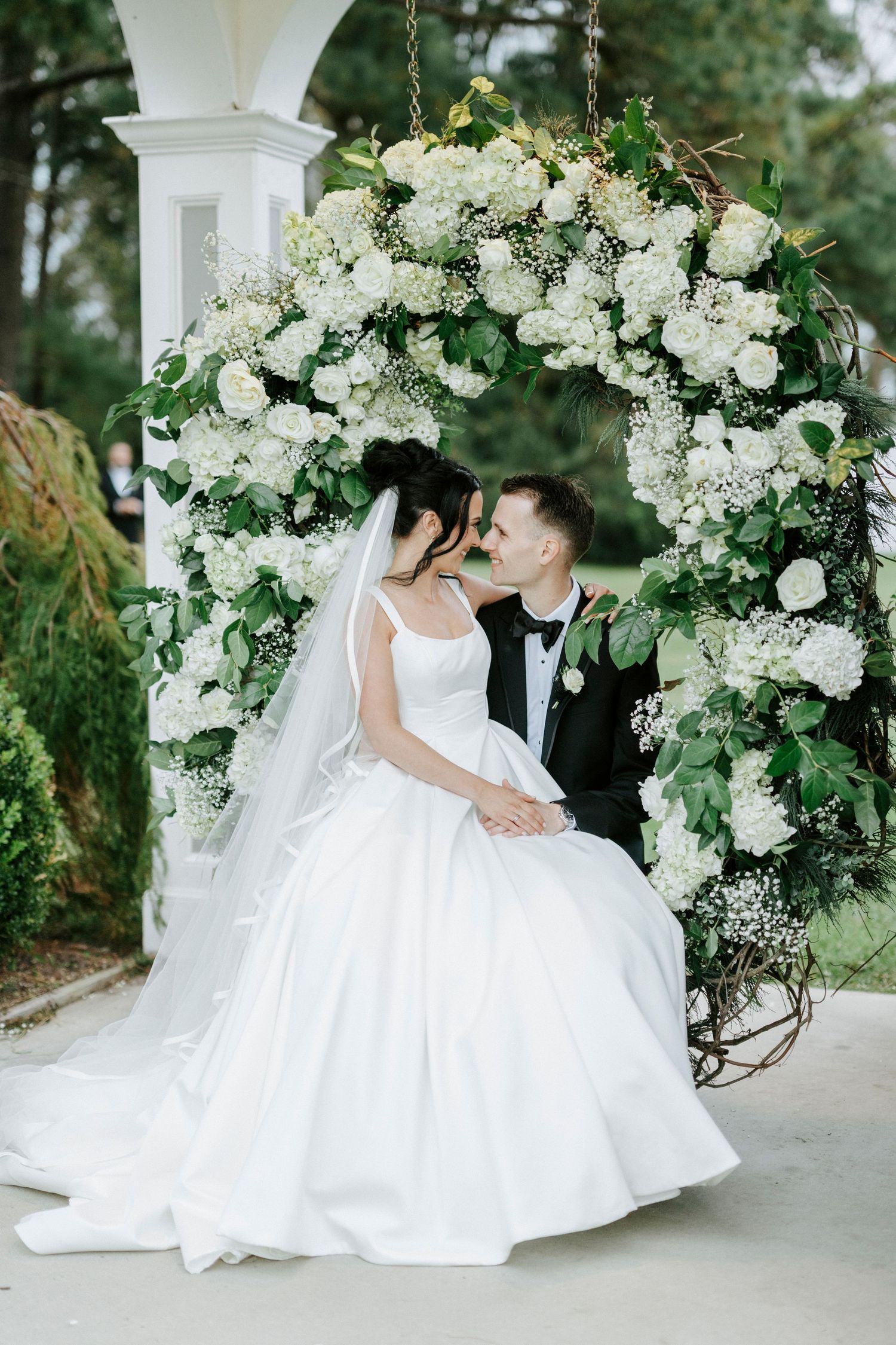 A romantic wedding scene with a floral arch adorned in white roses and greenery captures tender moments between newlyweds.