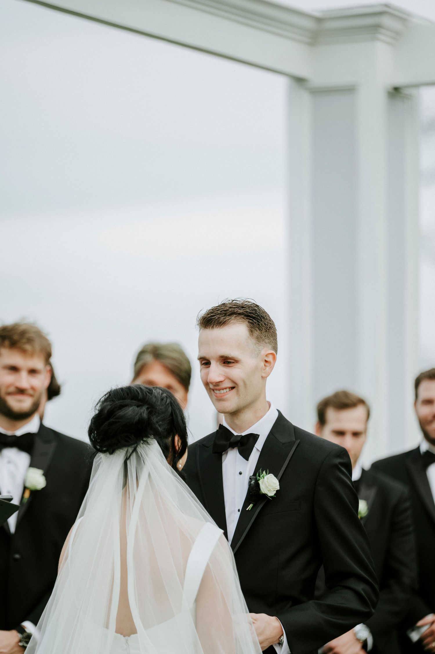 A wedding party in formal black tuxedos and white dresses gathers together in a bright white room.