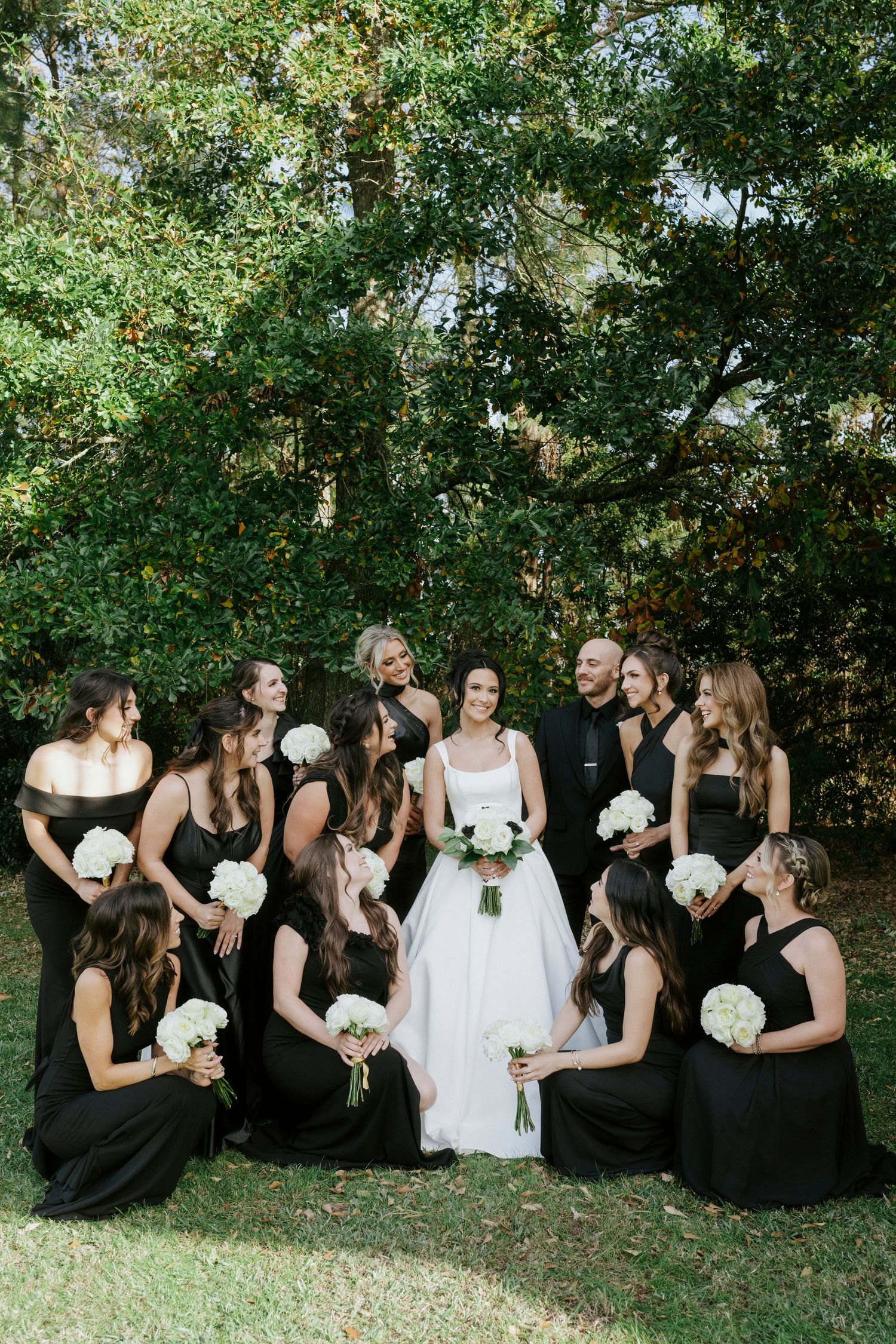 Bridesmaids in black dresses sit on grass with white bouquets while laughing with bride in white dress.