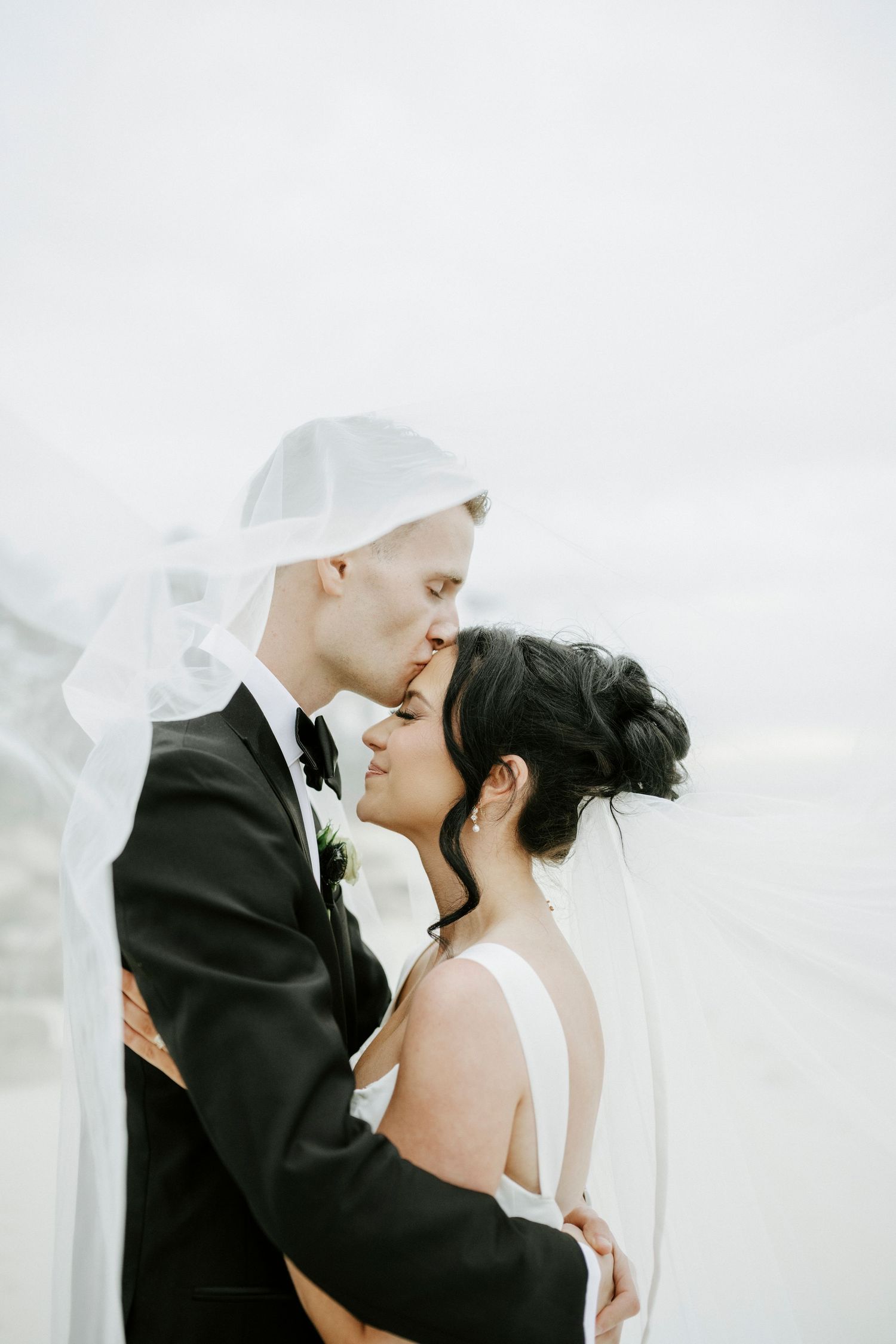 A tender moment between newlyweds as their veil flows in the breeze during an intimate outdoor wedding photoshoot.