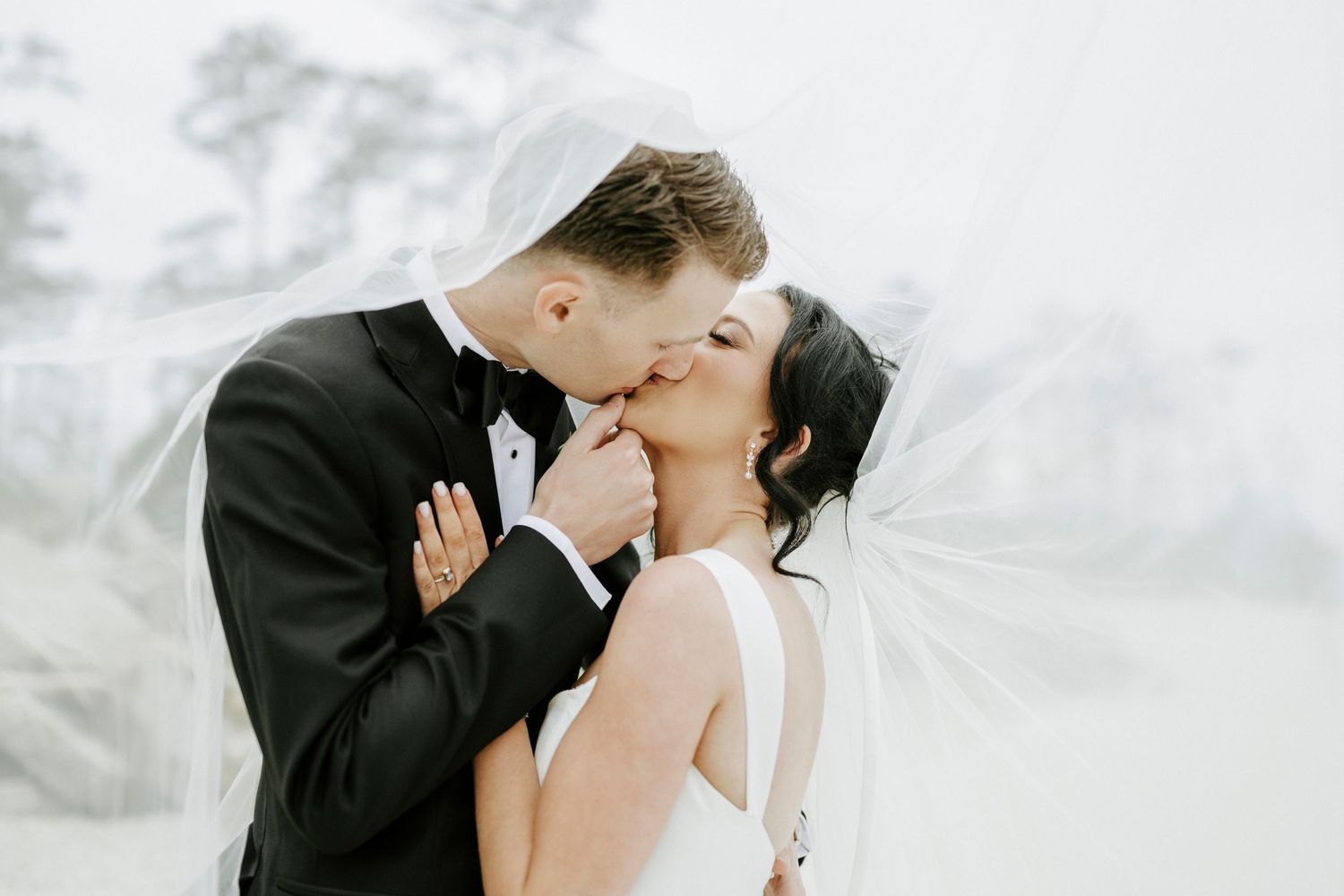 Elegant wedding couple in formal attire sharing romantic kisses on a bright beach.