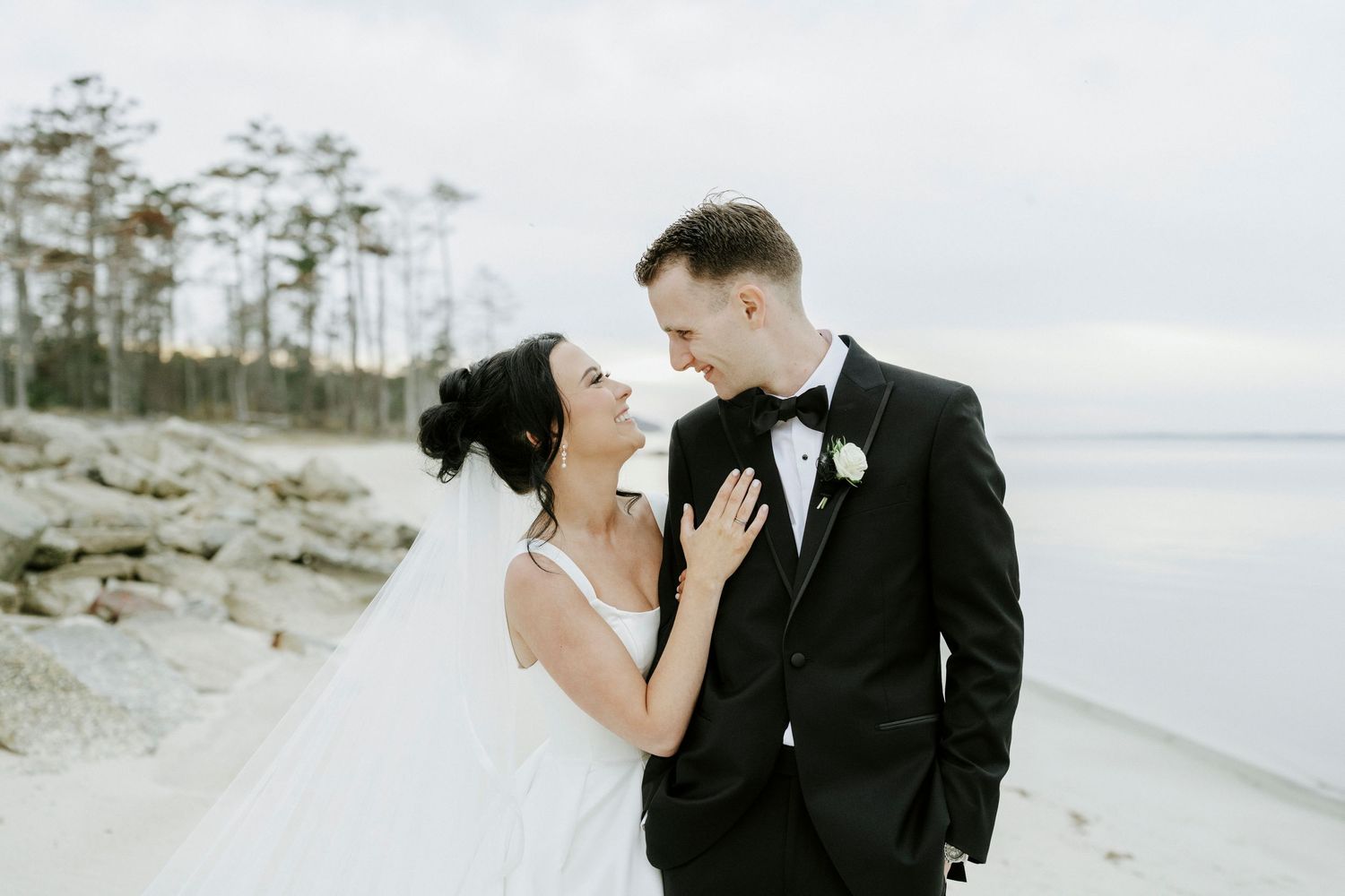 Bride in flowing white dress and groom in tuxedo share intimate moments on coastal shoreline.