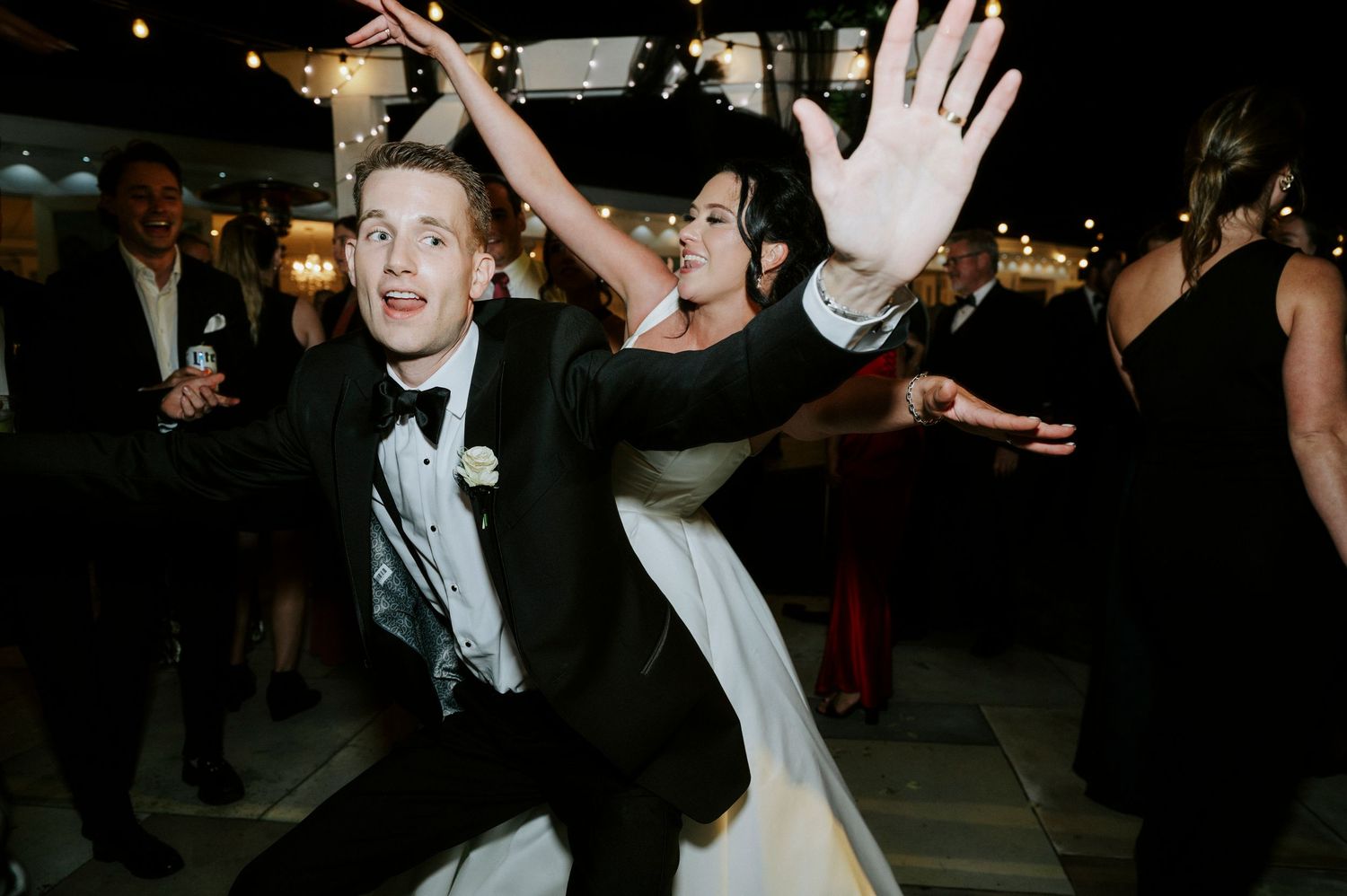 Wedding reception guests dancing and celebrating with hands raised on a dark dance floor.