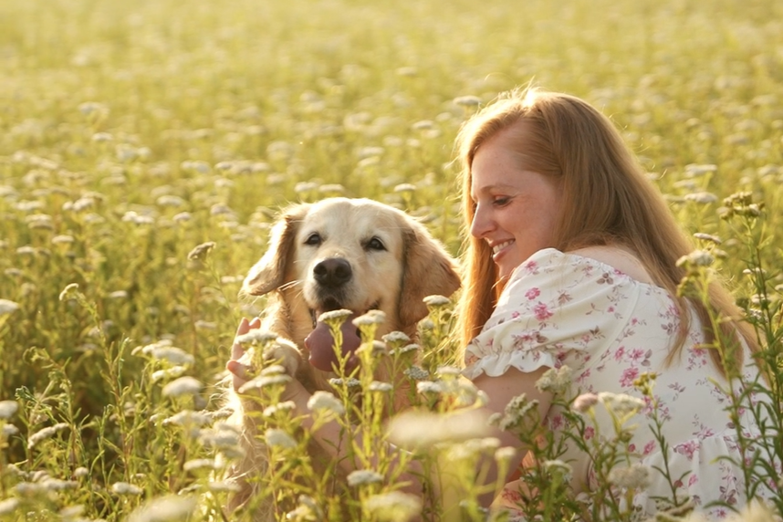 A smiling red-haired woman lies in a wildflower meadow beside her golden retriever in warm sunlight.