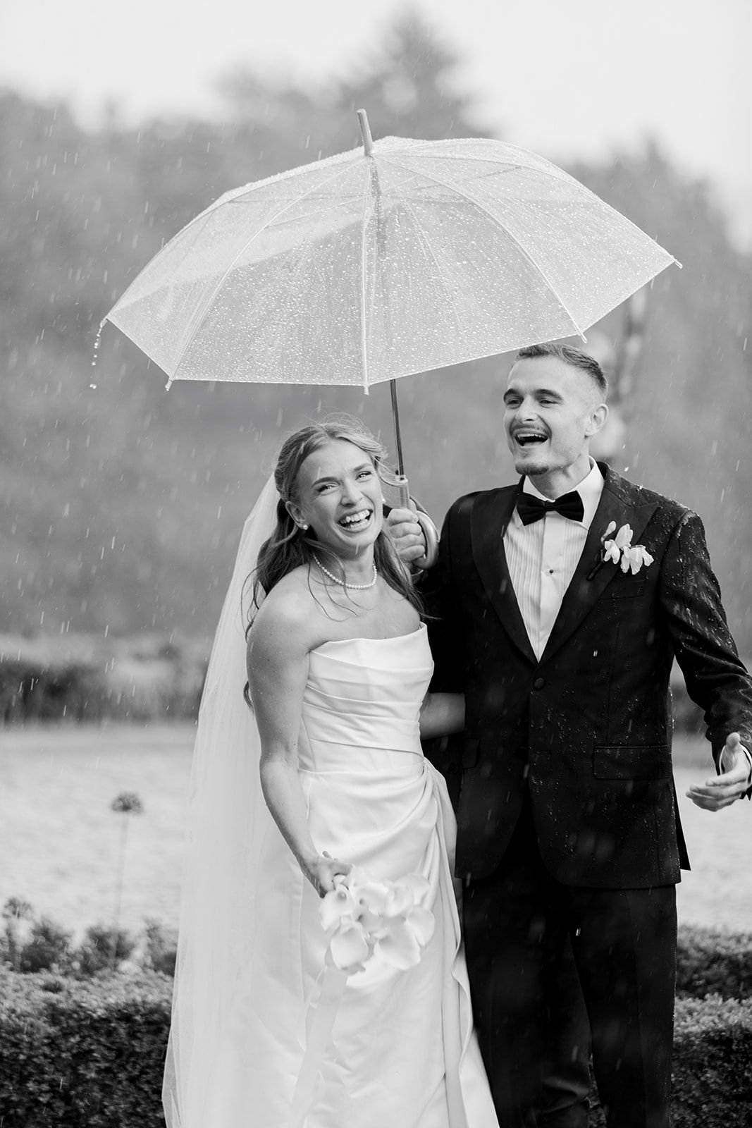 A couple shares an umbrella while walking in the rain on their wedding day, captured in black and white photography.