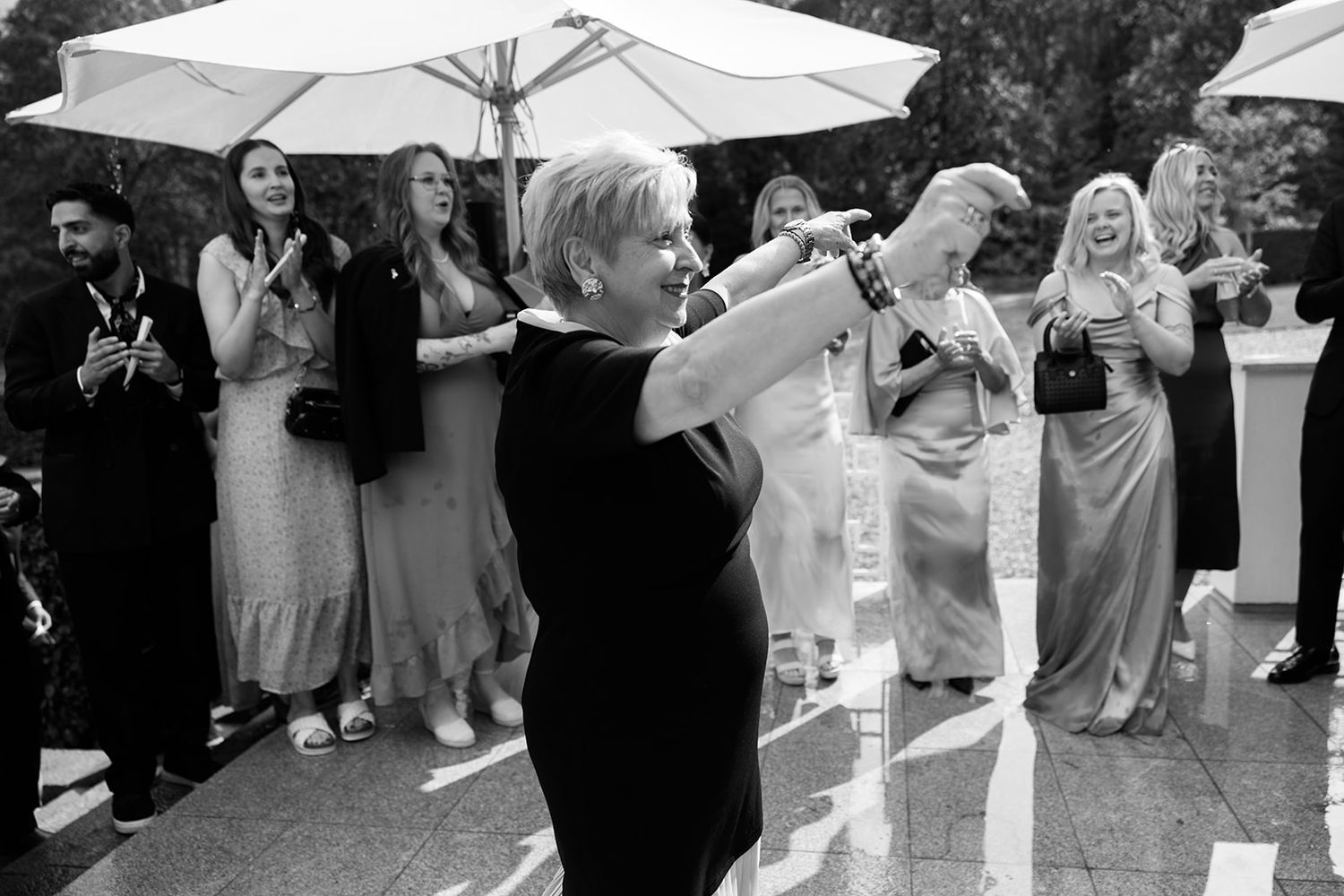 Wedding party celebrates joyfully under white umbrellas with groomsmen in black tuxedos and bridesmaids in colorful dresses.