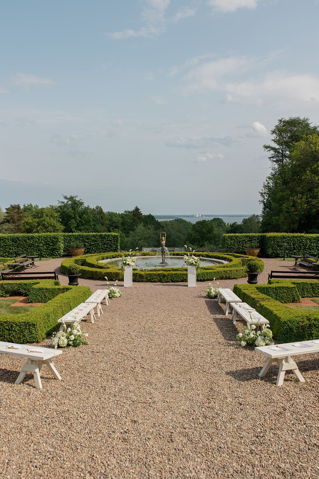 Outdoor wedding ceremony setup with white chairs and pedestals overlooking a scenic waterfront garden landscape.