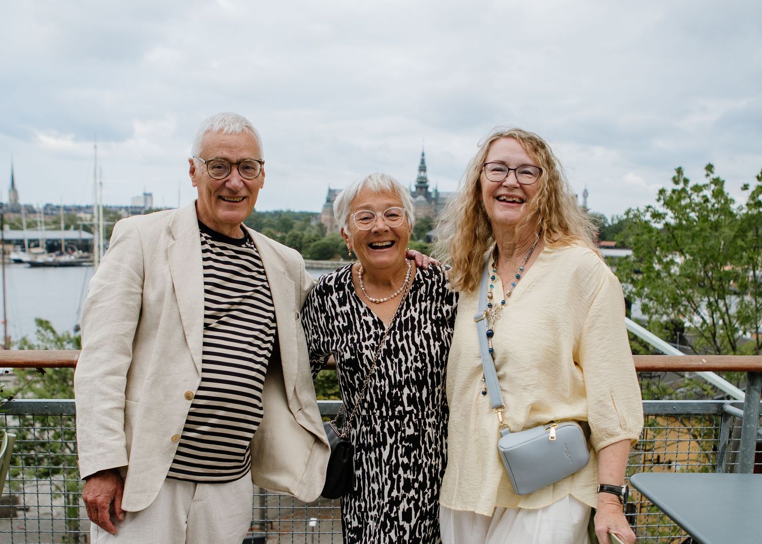 Three smiling seniors posing together outdoors on a waterfront terrace with Stockholm cityscape behind them.
