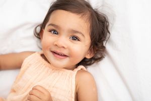 Baby with dark curly hair lies on white bedding while smiling at the camera.