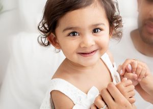 A toddler with curly hair and bright eyes smiles joyfully while playing with their hands.