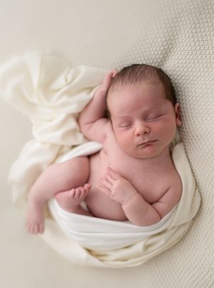 Peaceful newborn baby sleeping wrapped in a soft cream cloth on a white textured blanket.