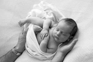 Black and white photo of a sleeping newborn baby wrapped in white cloth, cradled gently in adult hands.
