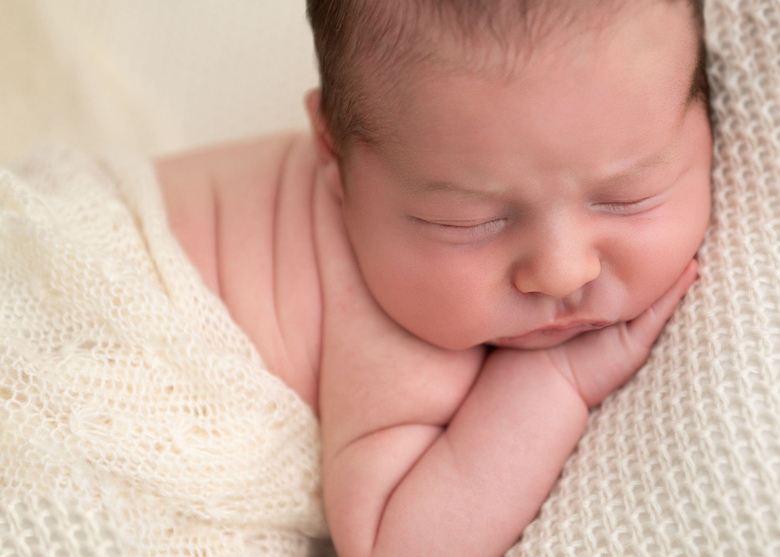 Peaceful newborn baby sleeping wrapped in a soft cream knit blanket, with chubby cheeks and tiny hands tucked under chin.