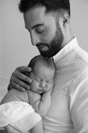 Black and white photo of a bearded father tenderly holding his sleeping newborn baby close to his chest.