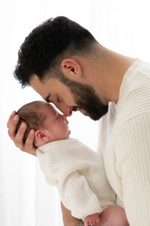 Bearded father tenderly touches foreheads with his swaddled newborn baby, both dressed in white.