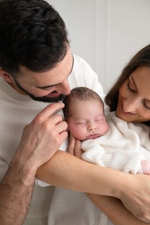 Loving parents gently cradling their sleeping newborn baby wrapped in a white knit blanket.