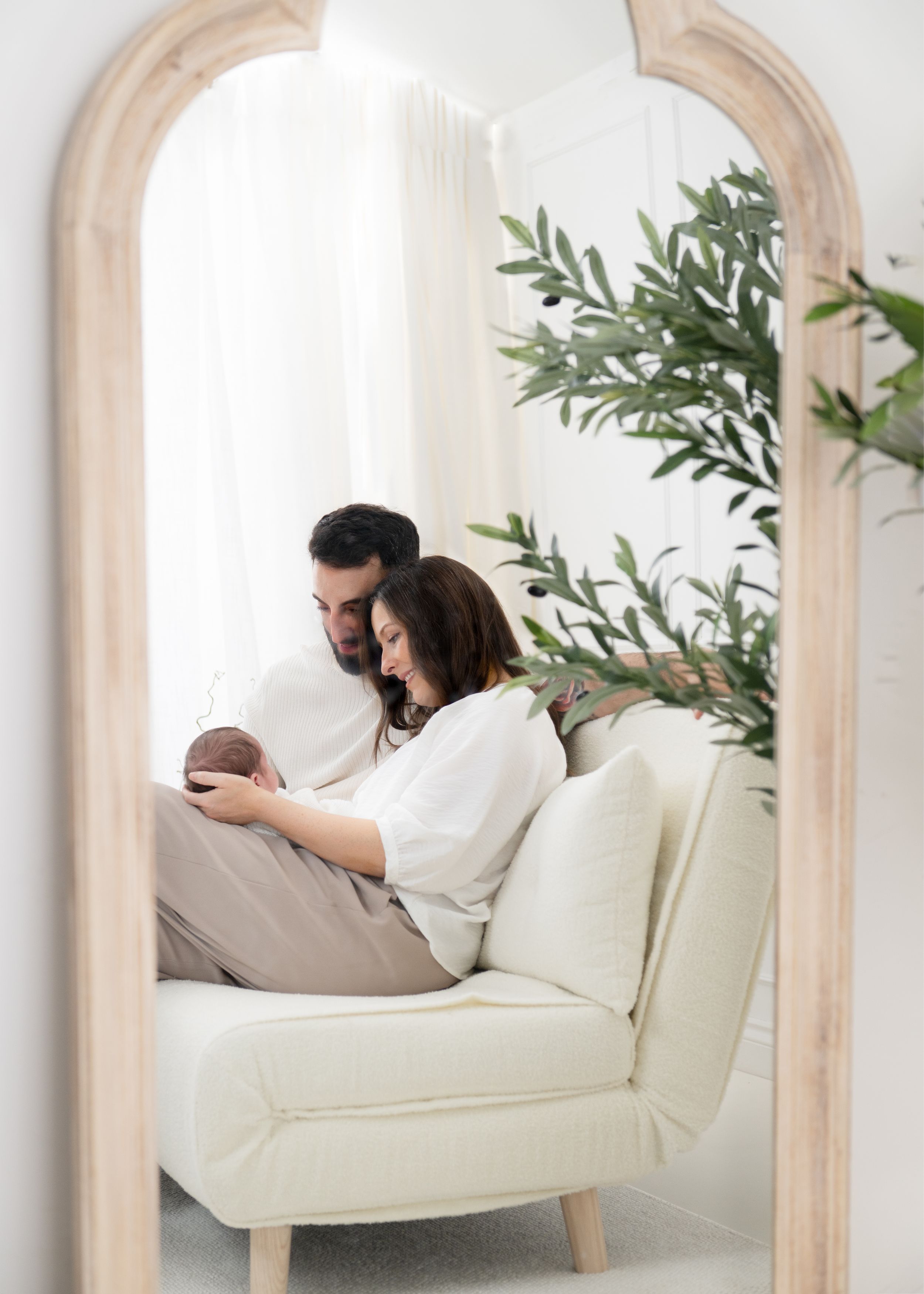 New parents tenderly holding their newborn baby together on a white sofa, reflected in an arched wooden mirror.
