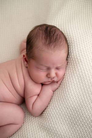 Peaceful newborn baby sleeping curled up on a soft cream knit blanket, with chubby cheeks and tiny hands tucked under chin.