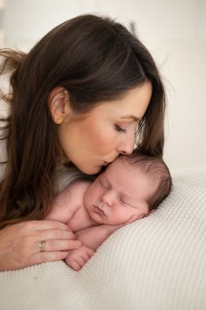 A mother with long brown hair gently kisses her sleeping newborn baby on a soft white blanket.