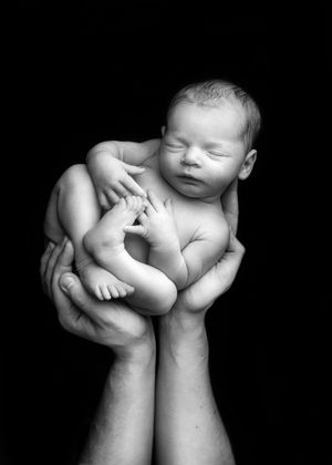 Black and white artistic portrait of a sleeping newborn baby cradled in gentle hands against a dark background.