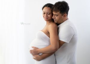 A couple wearing white embraces tenderly in a bright studio setting during a maternity photoshoot.