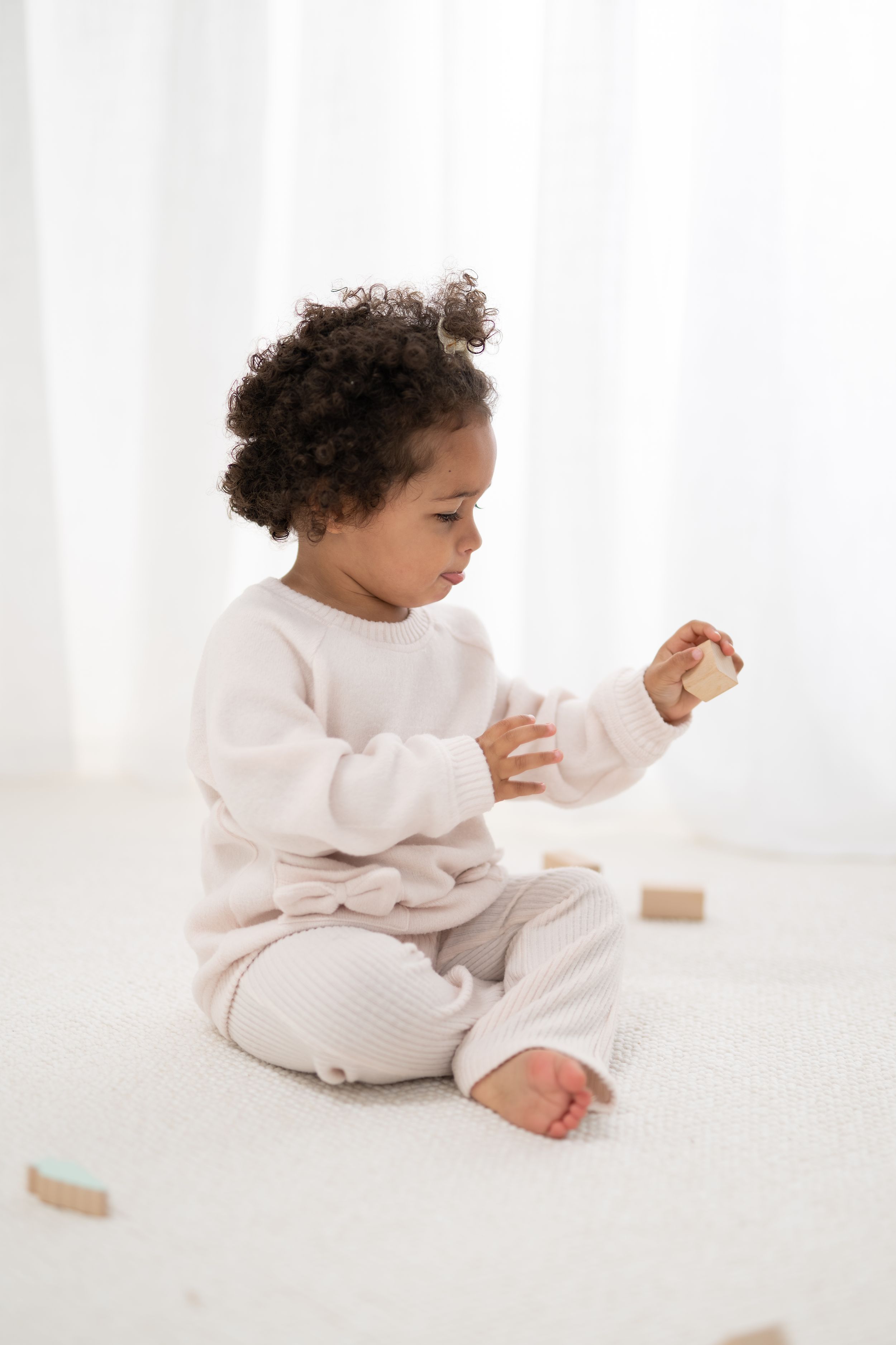 A toddler in a cozy white sweater and pants sits on a light floor while playing with small toys.